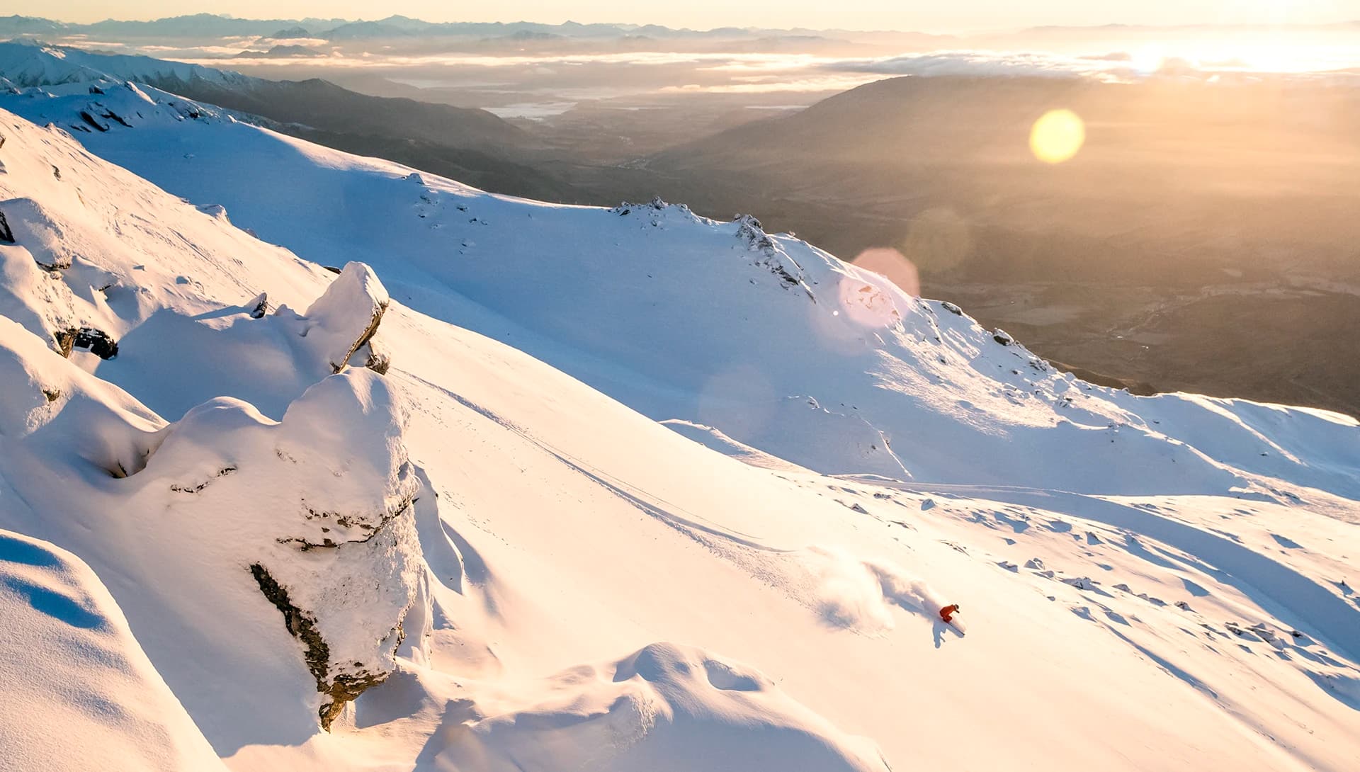 A lone skier in red glides down a vast, sunlit snow-covered mountain. The golden sunset and distant peaks create a serene backdrop.