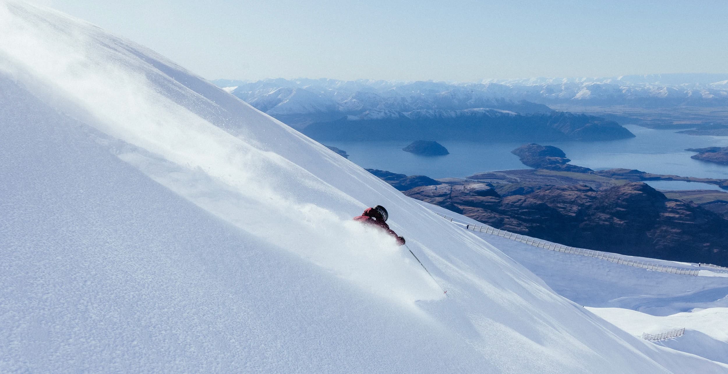 A skier carves down a steep, snowy slope with powder flying, while a vast lake and snow-capped mountains stretch across the background.