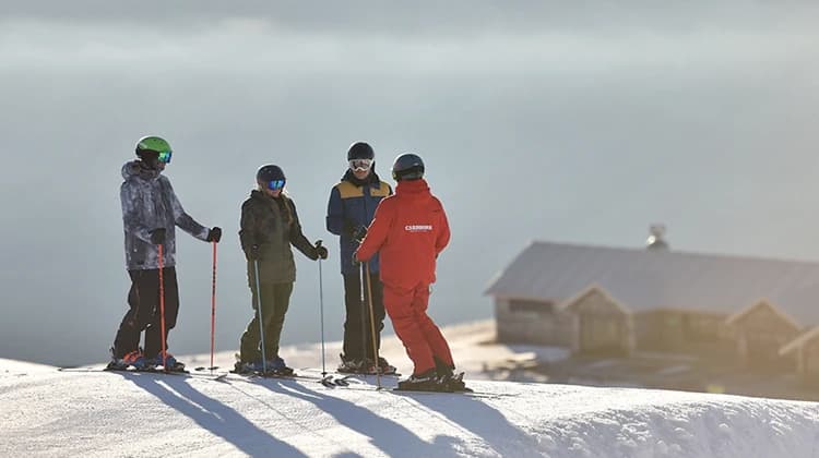 A ski instructor in a red jacket talks with three skiers on a snowy slope. The morning light creates long shadows, with a mountain lodge visible in the background under a serene sky.