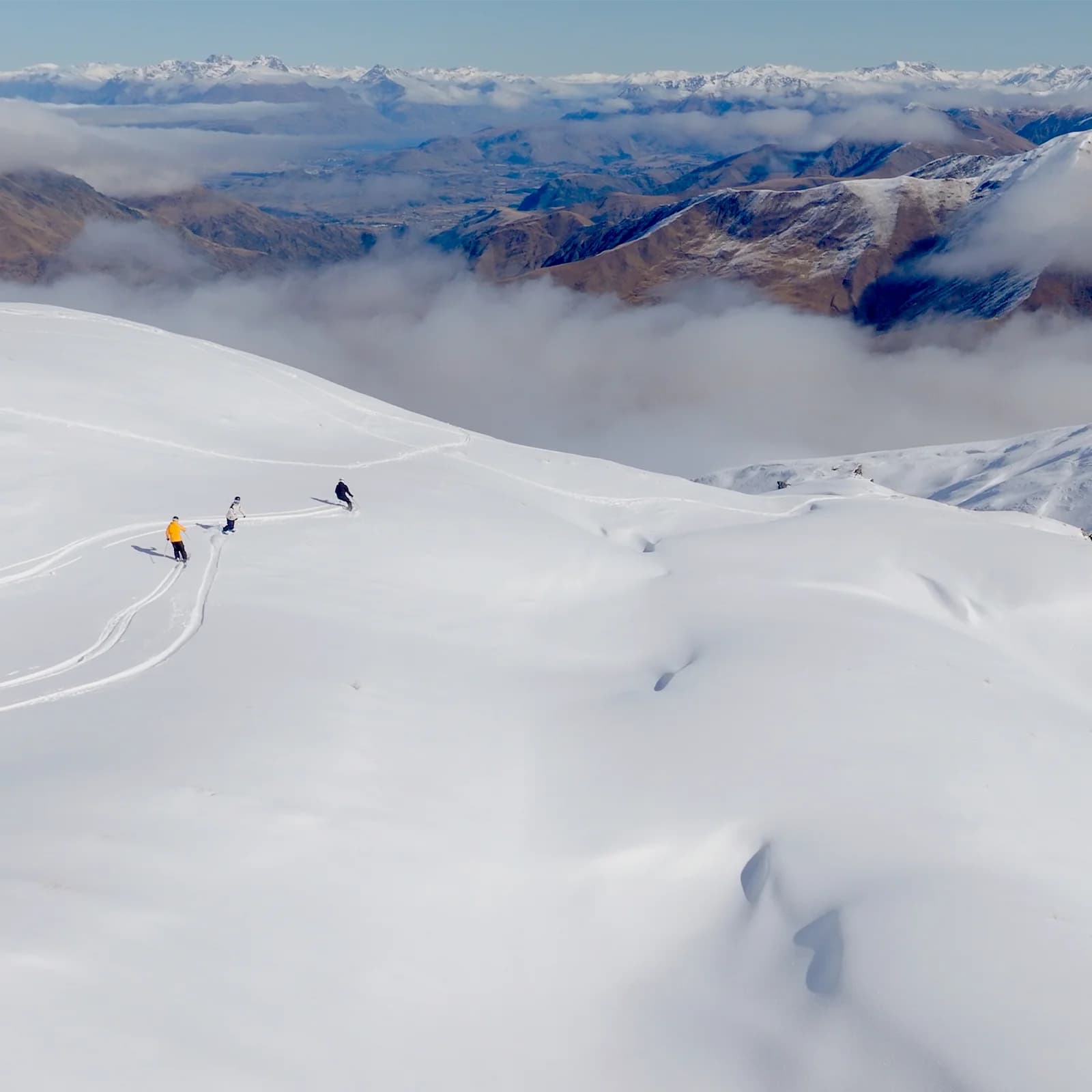 Three skiers glide down a vast, untouched snowfield, leaving tracks behind them, with a backdrop of majestic mountains and low-hanging clouds filling the valleys below.