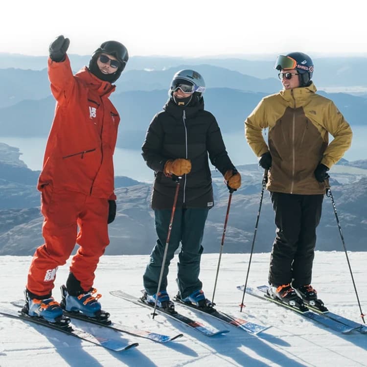 Two people taking a ski lesson at the top of Treble Cone Ski Resort, New Zealand, with instructors guiding them on snow-covered slopes and a panoramic view of surrounding mountains.