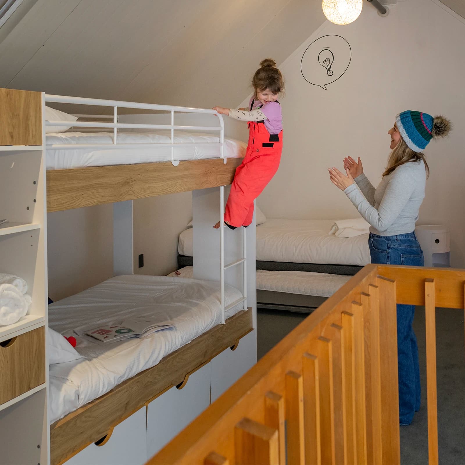 A child dressed in bright coloured ski gear climbs up the ladder of bunk beds in a cosy room. A woman, also dressed in snow gear, stands behind the girl smiling.