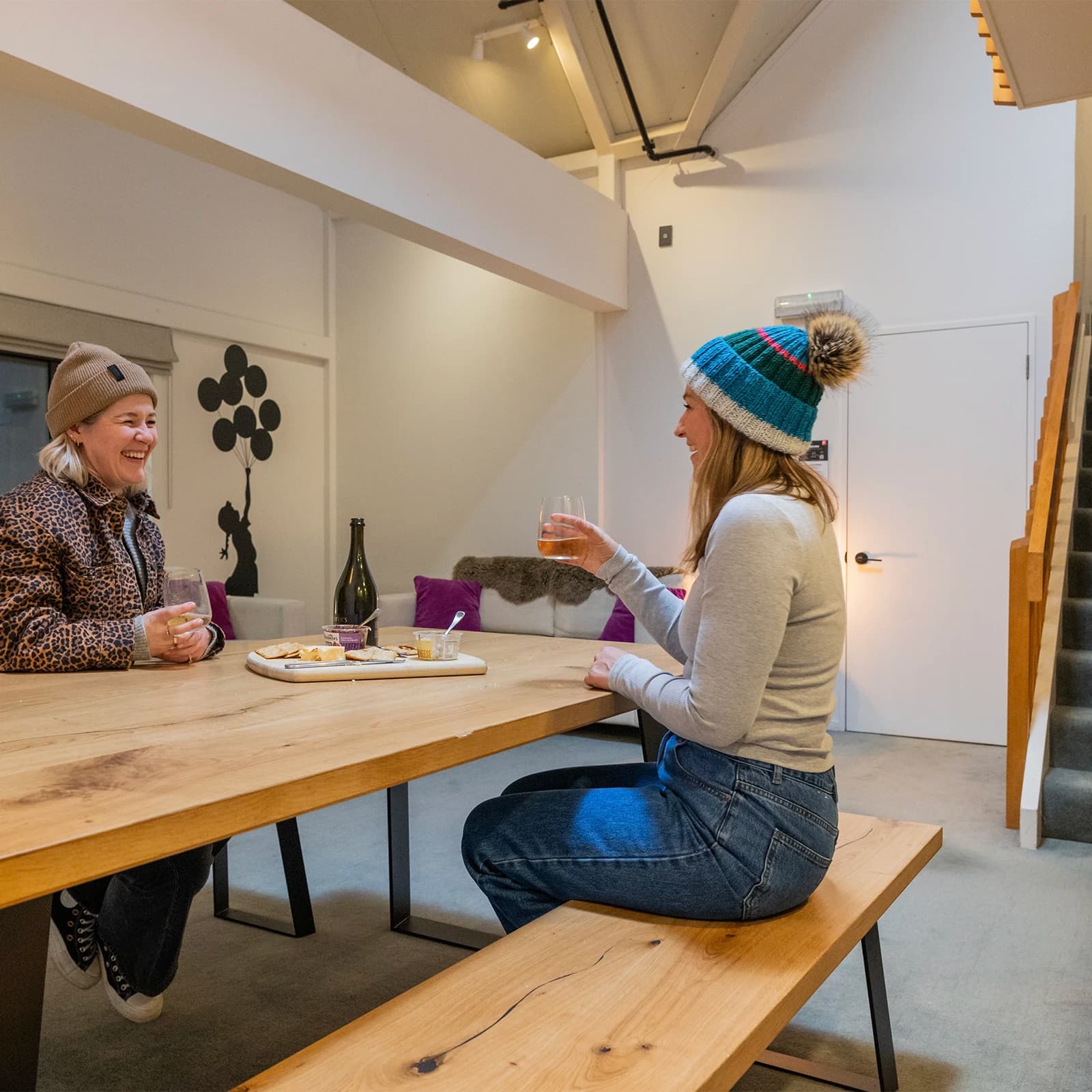 Two women enjoy a cheese board and bottle of wine in a cosy apartment.