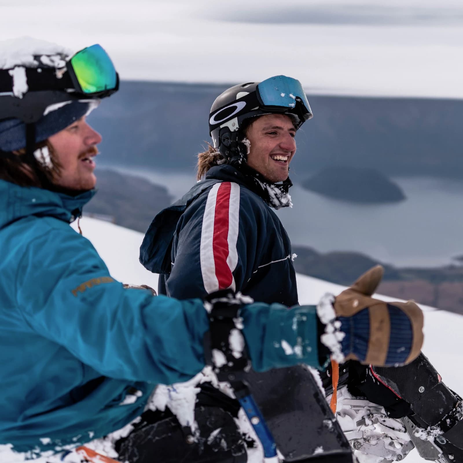 Two smiling athletes in sit skis at the Treble Cone Saddle, with views of Lake Wanaka in the background. 