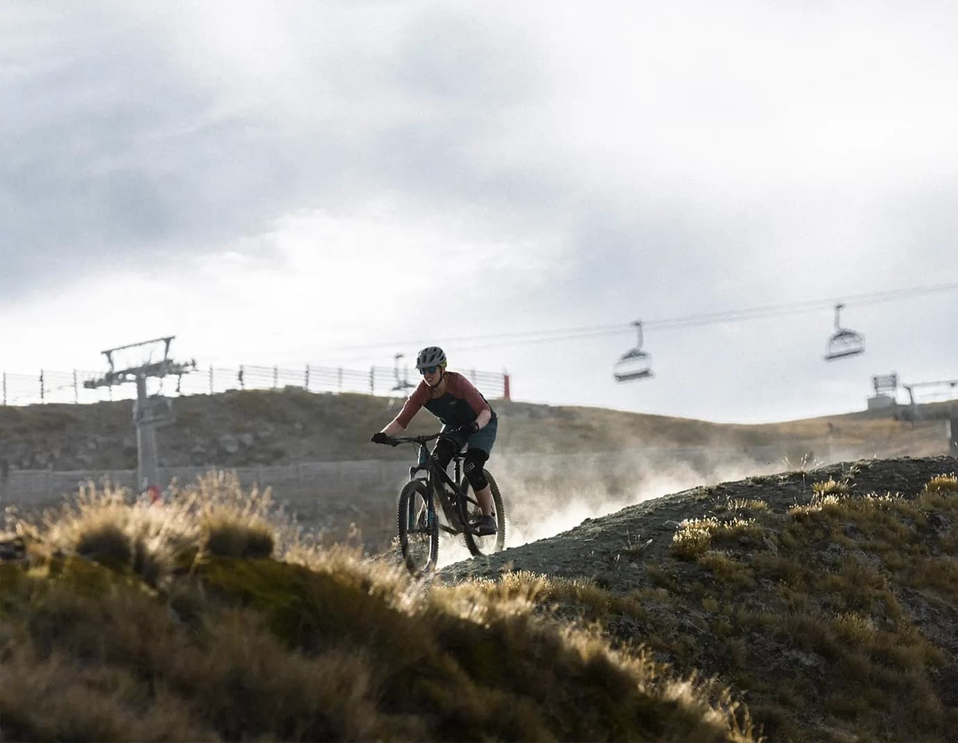 A mountain biker riding on a trail, with ski lifts in the background.