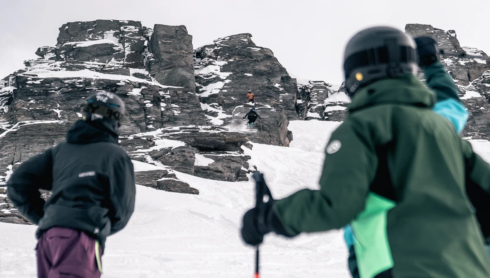 Two skiers in the foreground watch a third skier performing a jump from a rocky outcrop in a snowy landscape. The skier in the air is mid-jump. 