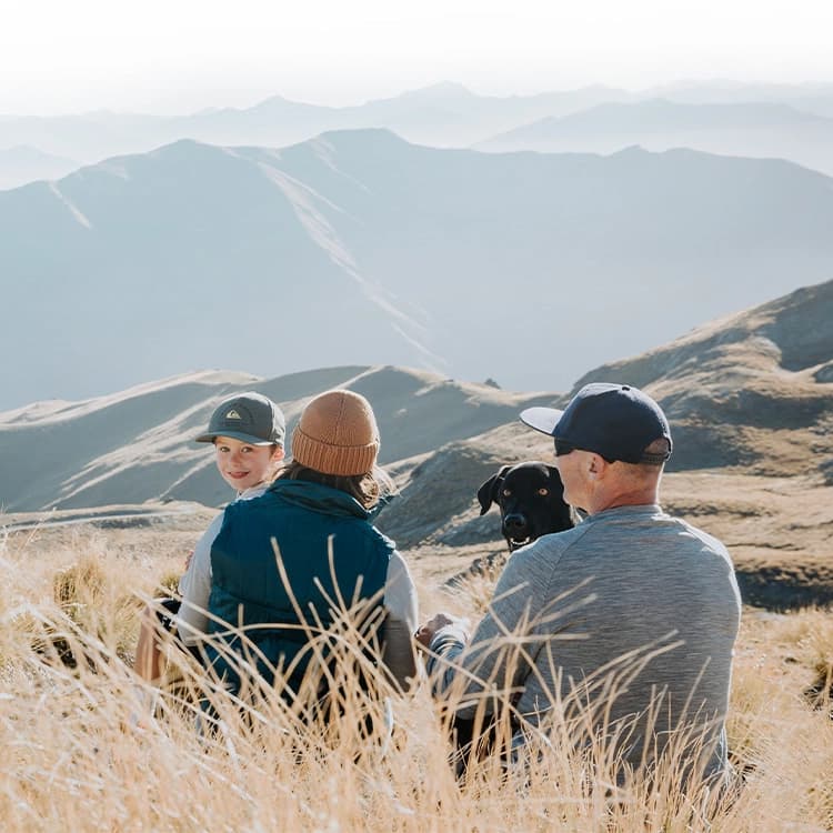 A family of two adults, one child, and a black dog sit amidst tall, golden grass on a mountainous terrain. The background features a panoramic view of softly lit mountain ranges under a clear sky. The child and the dog are facing the camera, while the adults are turned towards each other, enjoying the serene landscape.