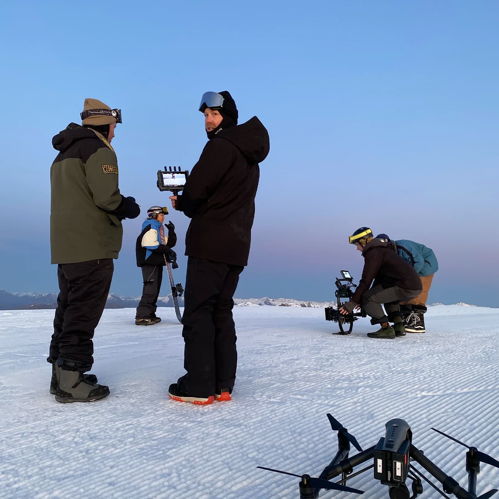 A film crew set up cameras an equipment on a snowy mountain top on a clear blue morning.