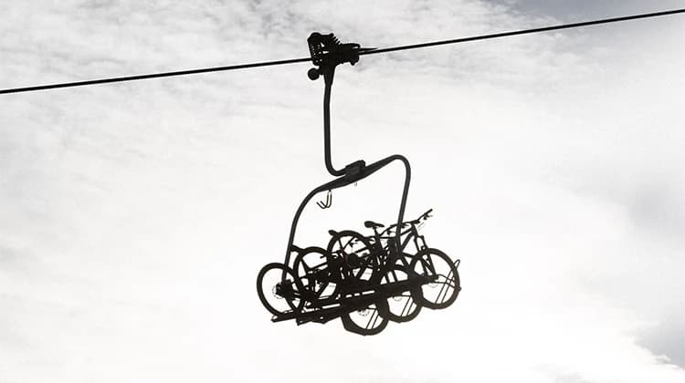 A chairlift silhouetted against a cloudy sky carries several mountain bikes. The bikes are secured on a specialized rack attached beneath the seat of the lift.