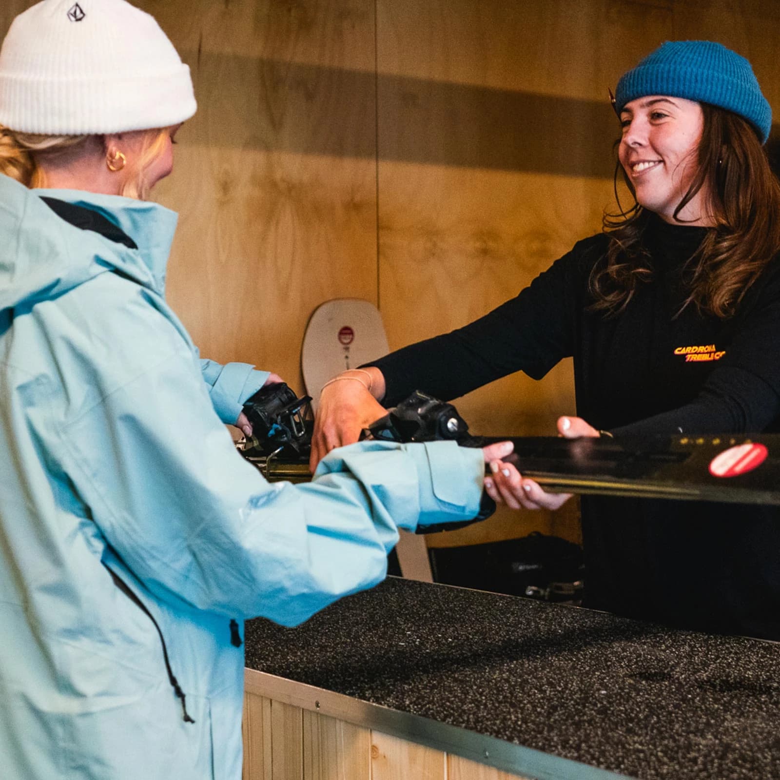 An indoor scene at a ski rental shop with a female technician handing skis across the desk to a customer. The technician is wearing a black long-sleeve shirt with the "Cardrona Rentals" logo. The customer, partially visible, is wearing a light blue jacket and a white beanie.