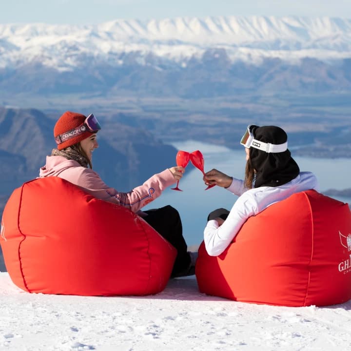 Two skiers recline in red branded beanbags on a snowy summit, toasting with red wine glasses overlooking a lake and mountains.