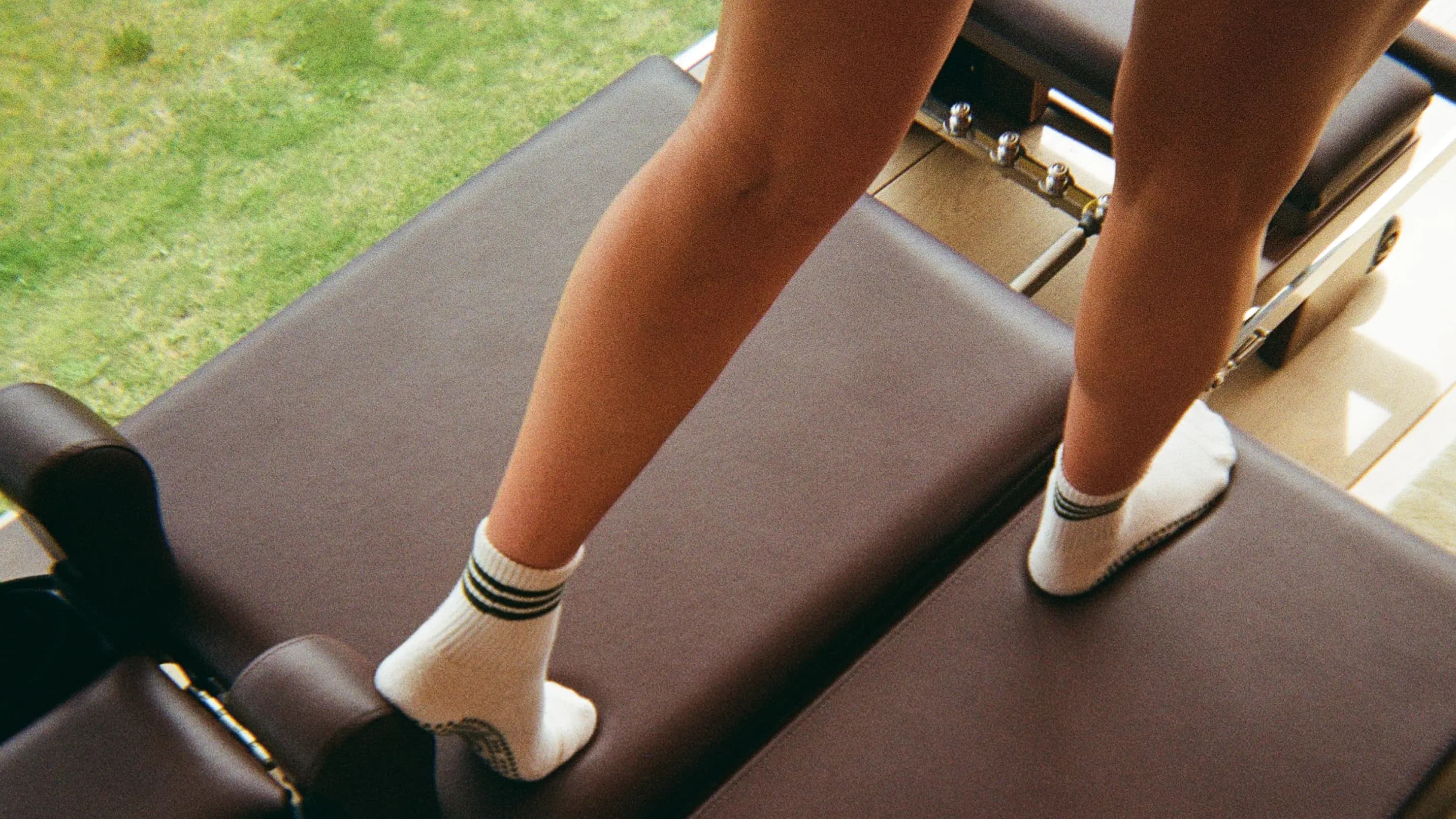 A person standing on a Pilates reformer machine in white grip socks, with green grass visible through the window behind them.
