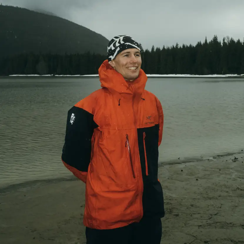 Headshot of NZ skier Craig Murray smiling by a lake on a moody day, wearing an orange jacket and black beanie.