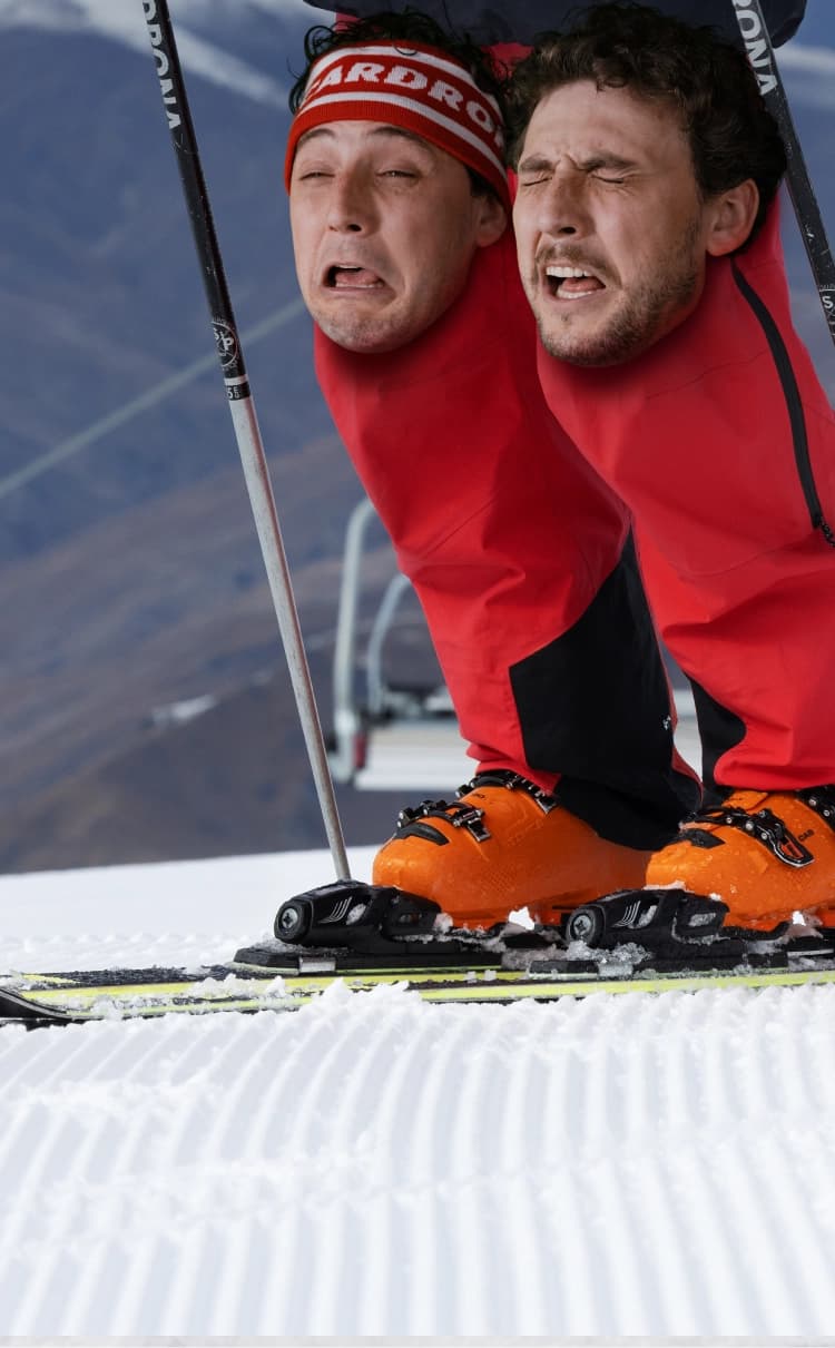 Two faces over the knees of a skier on a groomed slope with mountain scenery in the background.