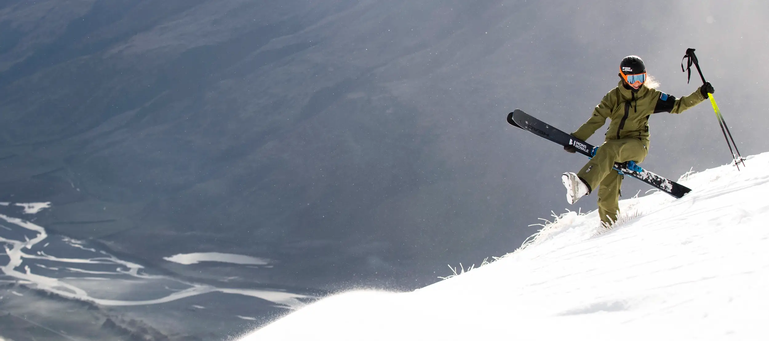 A person in ski gear joyfully hikes up a snowy mountain slope carrying skis and poles, with a dramatic alpine valley and braided river in the background.
