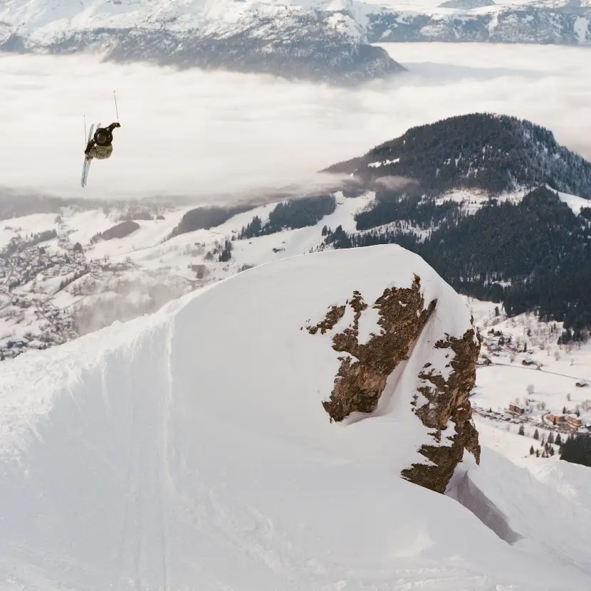 New Zealand skier Ben Richards launching off a massive cliff face high above an alpine valley, with clouds rolling through the mountains below.