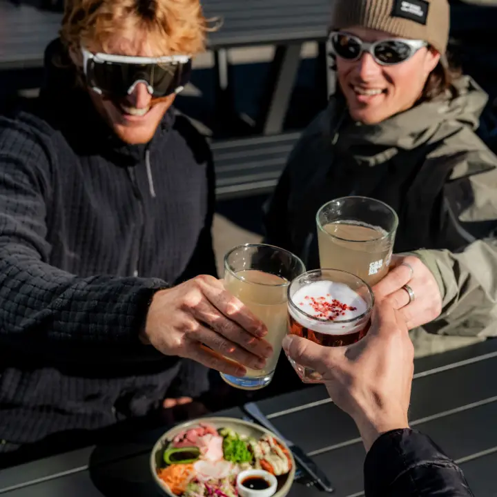 Three people in ski gear raising drinks in a sunny outdoor bar setting.