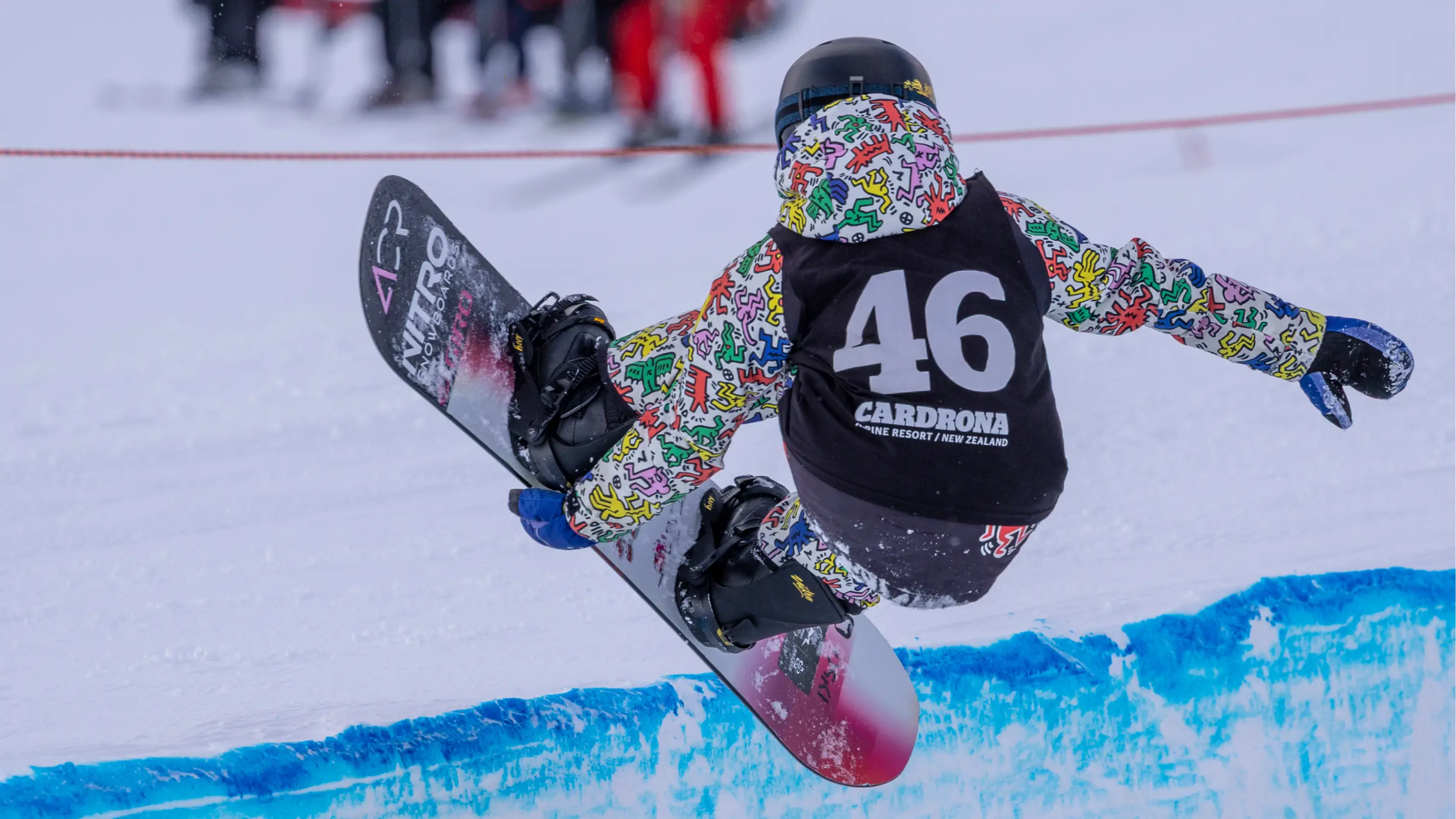 A snowboarder wearing bib 46 catches air out of a halfpipe at Cardrona Alpine Resort, with a chairlift and riders visible in the background.