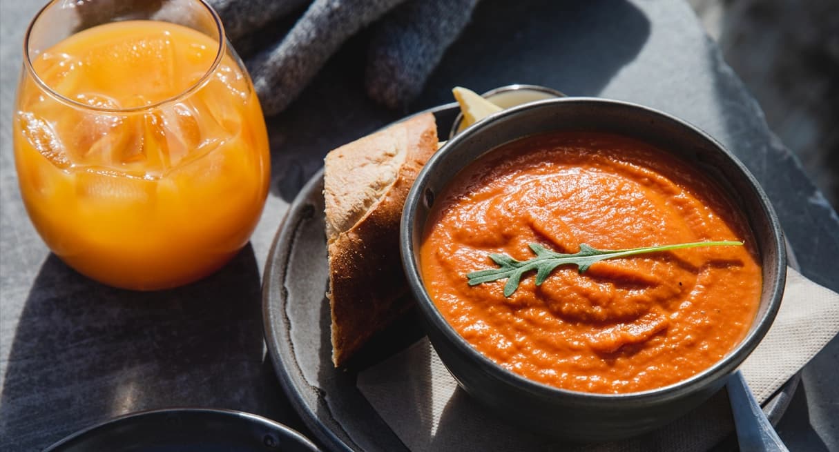 Tomato soup with bread, a glass of orange juice, and a chocolate dessert on a dark table in sunlight.