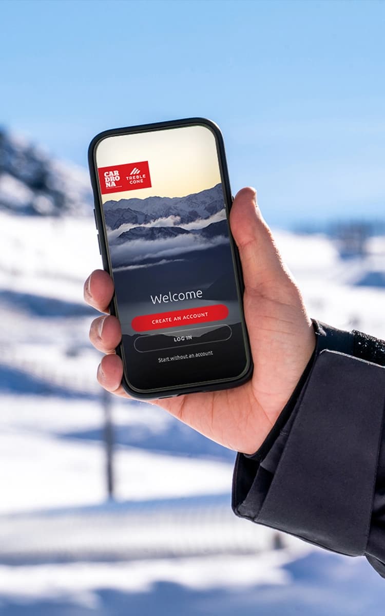 A hand holding a smartphone displaying the Cardrona and Treble Cone ski resort app welcome screen, with a snowy mountain landscape in the background.