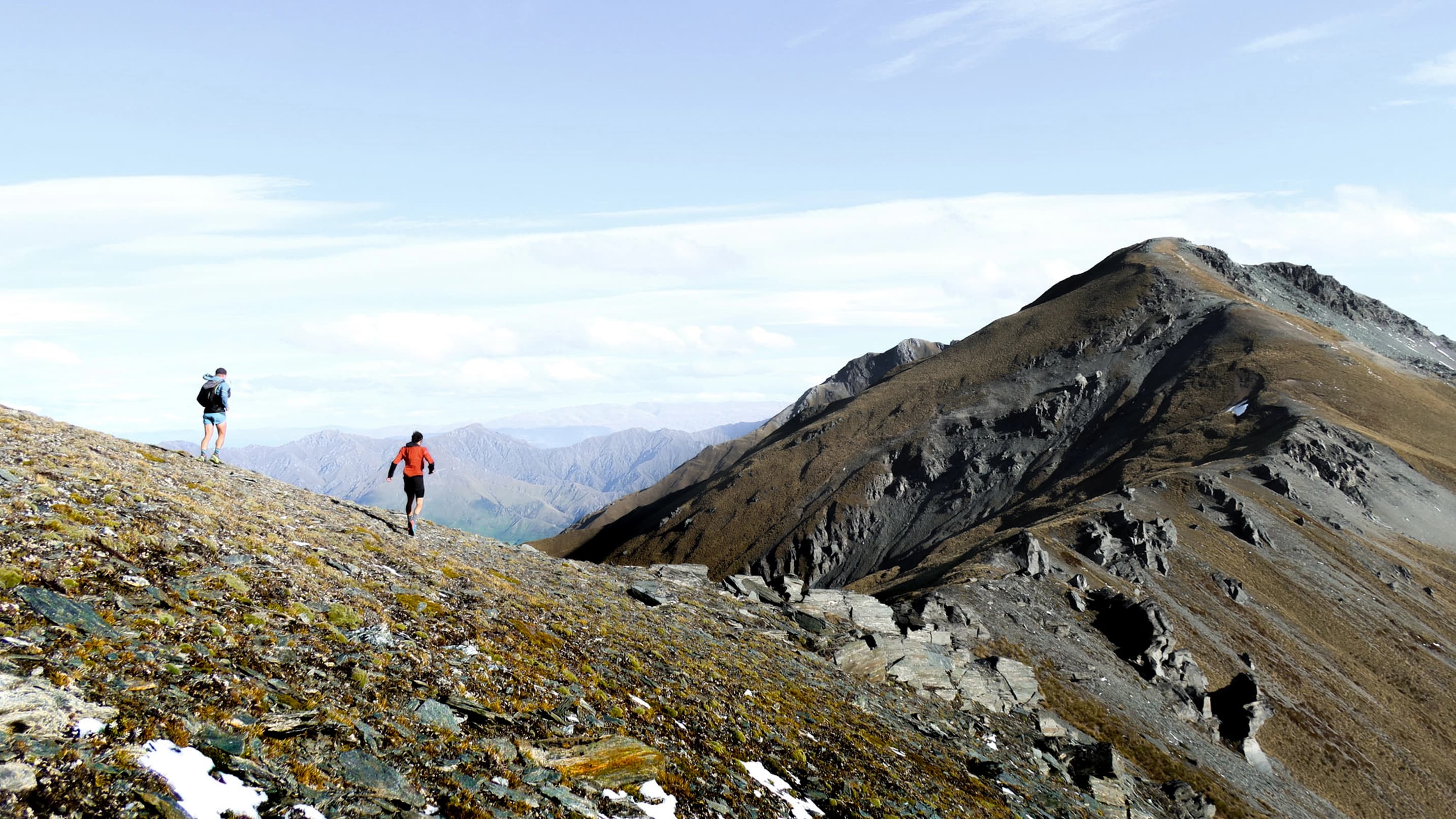 Two people running along the ridge of a rocky mountain on a clear blue skies day.