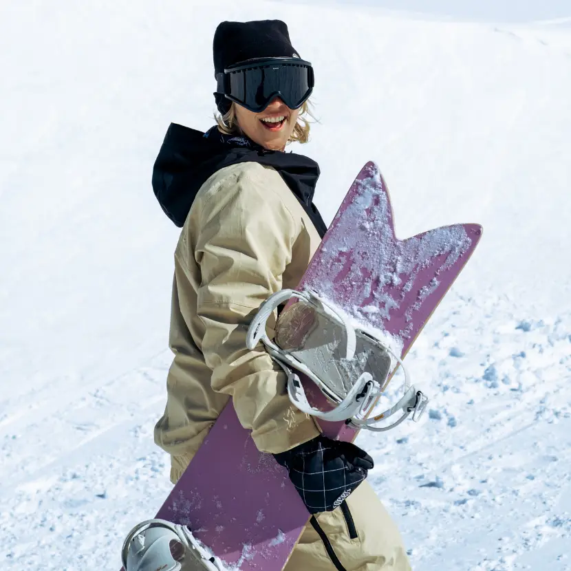 A smiling snowboarder carries their purple snowboard on a snowy mountain.