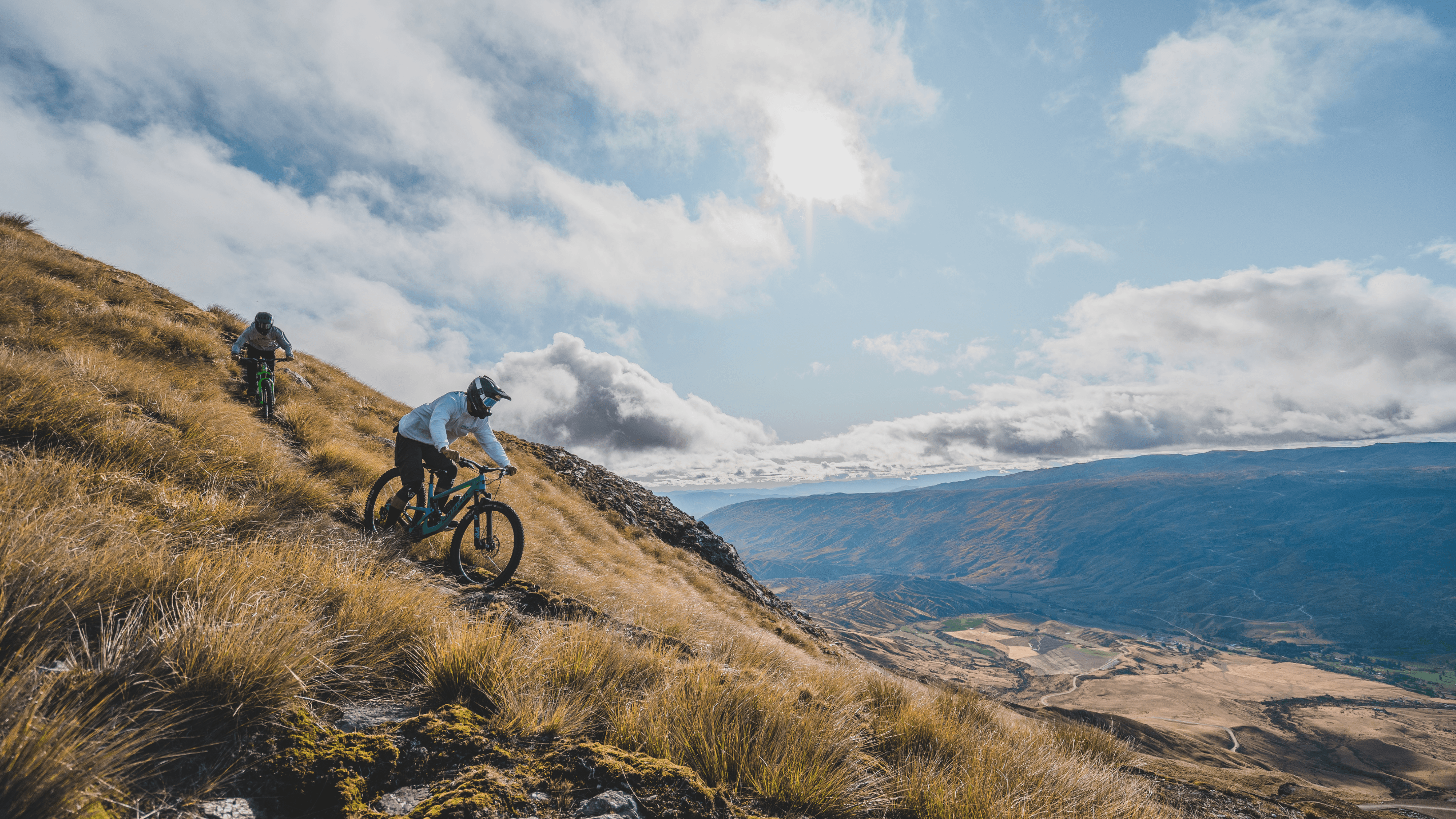 Two mountain bikers descending a grassy hill in full face helmets.