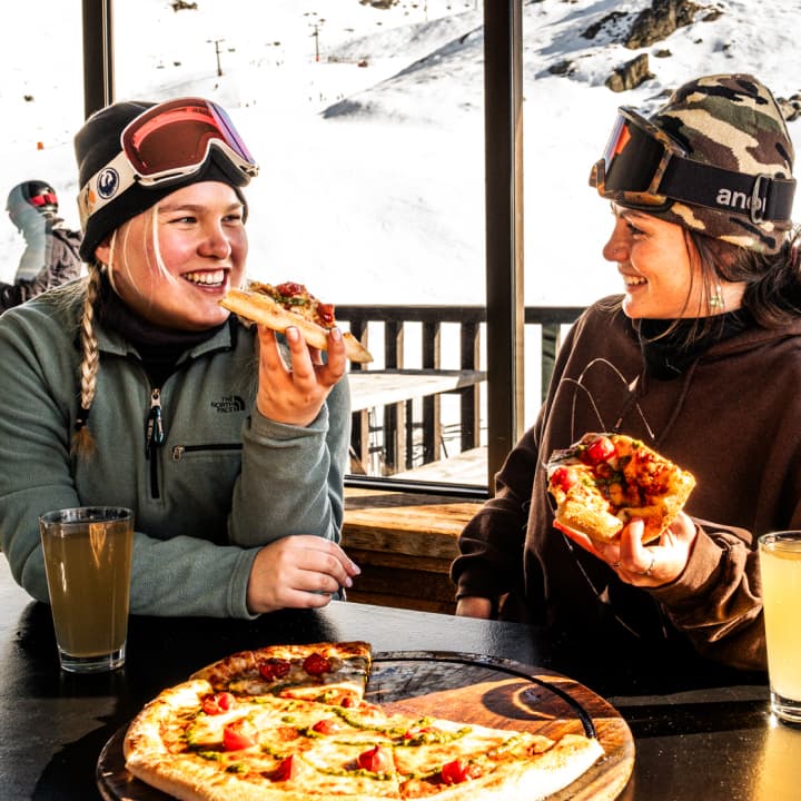Two smiling skiers eat pizza slices at an indoor table with snowy mountain views through the window behind them.