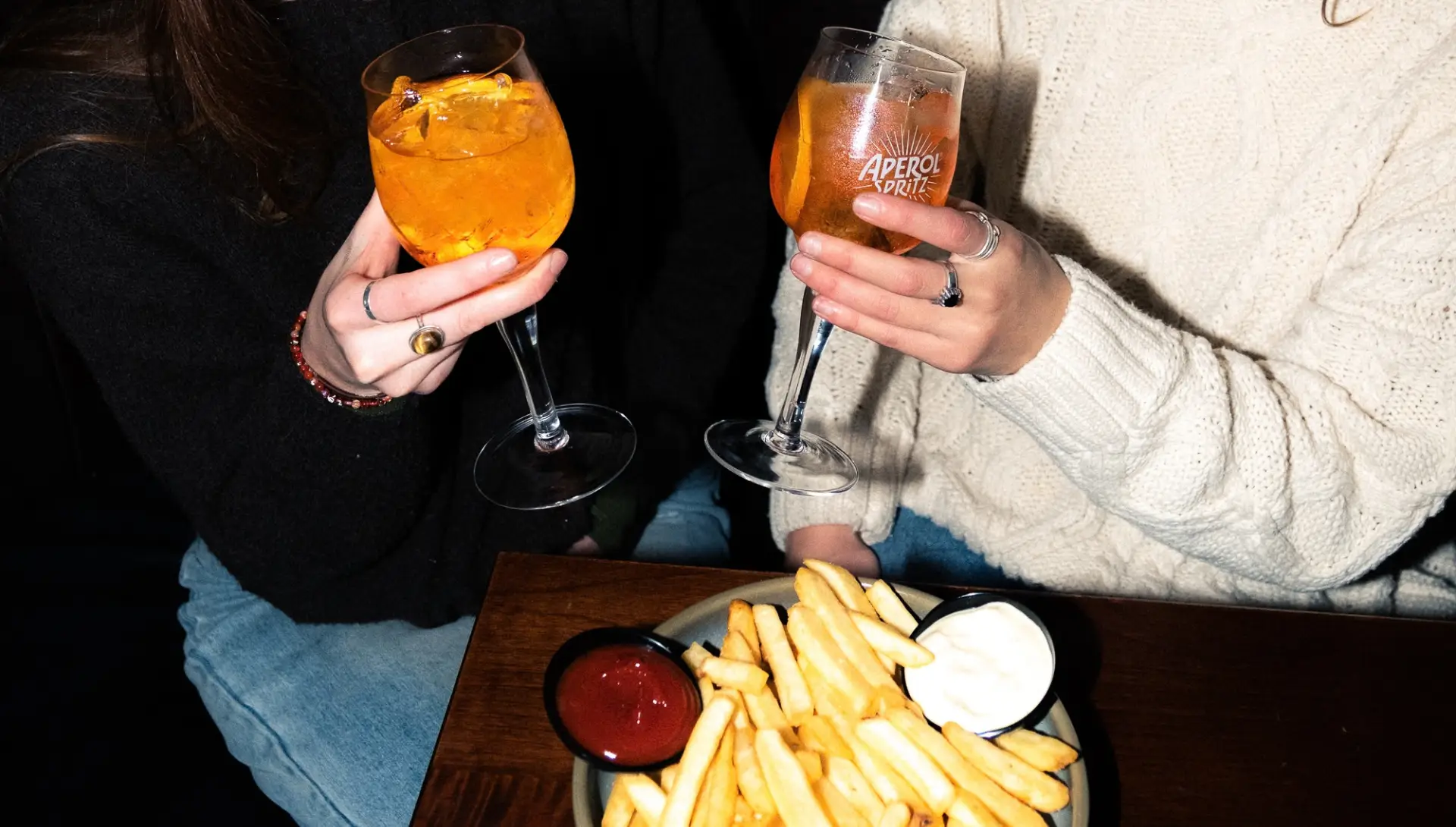 Two friends sitting side by side have an aperol spritz each and a plate of fries to share.
