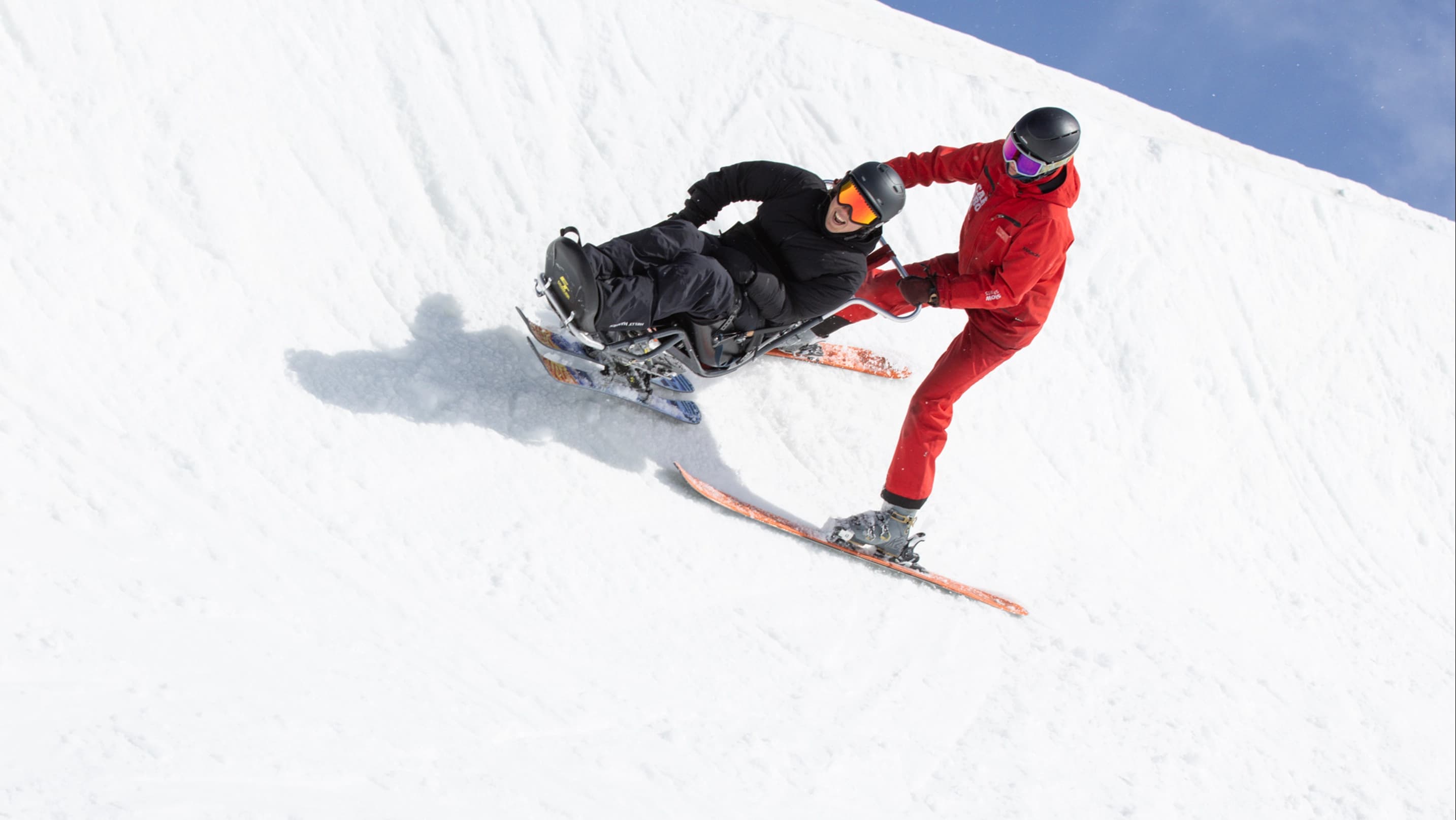 A sit-ski rider is guided downhill by an instructor in red on a snowy slope.