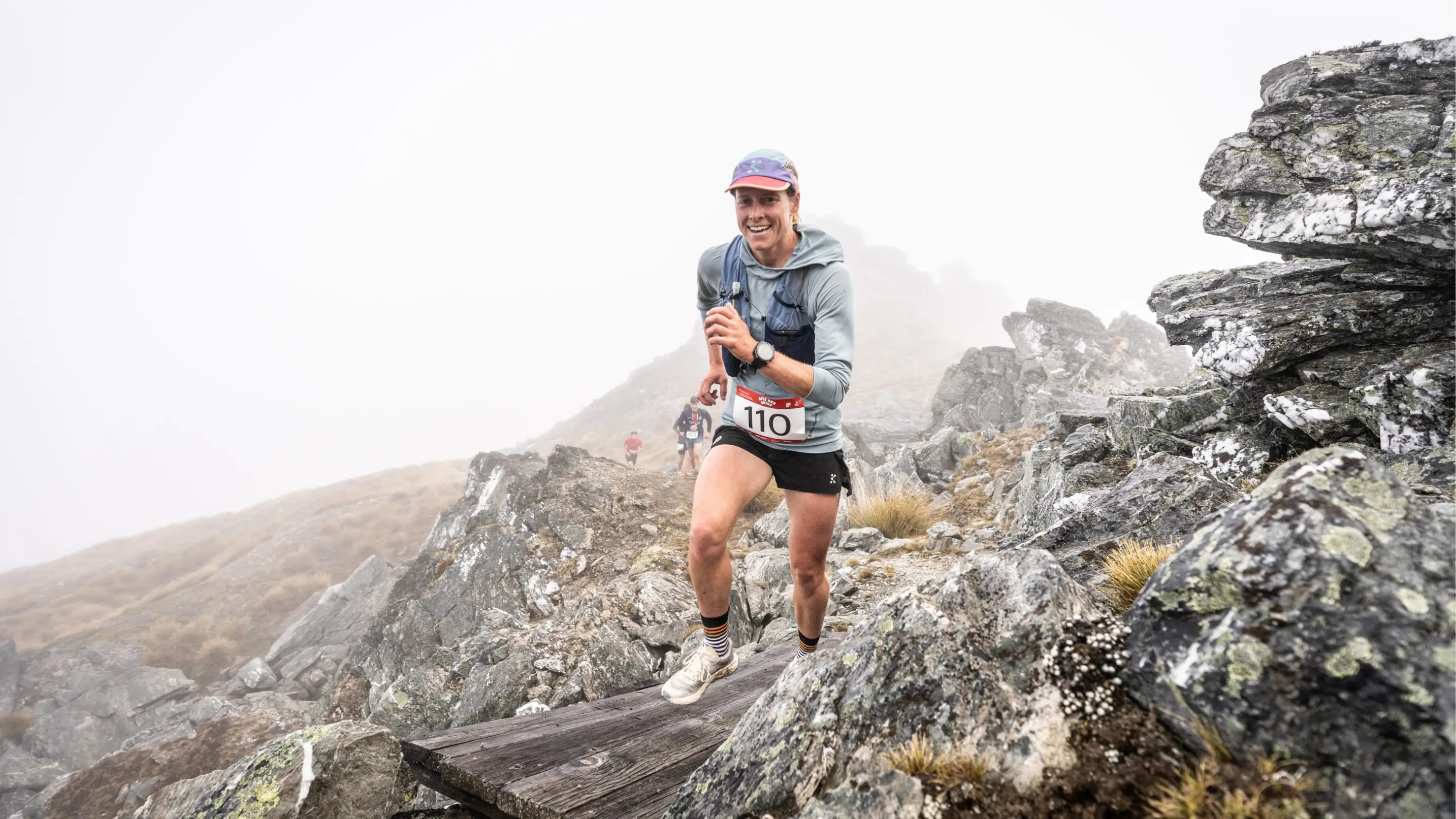 A trail runner jogs across a plank of wood on a rocky mountain trail.