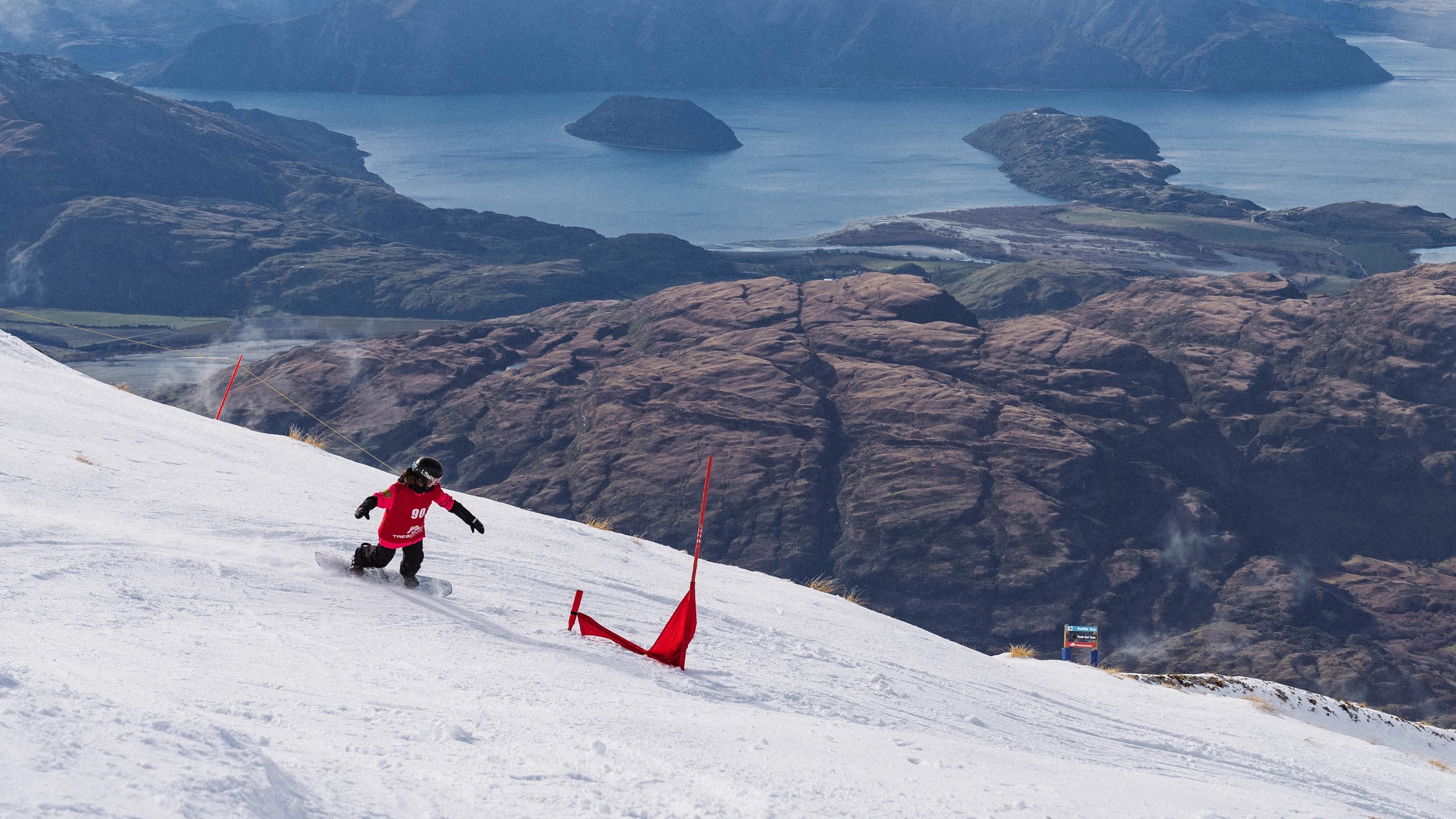 Snowboarder racing through a banked slalom course with sweeping lake views below.