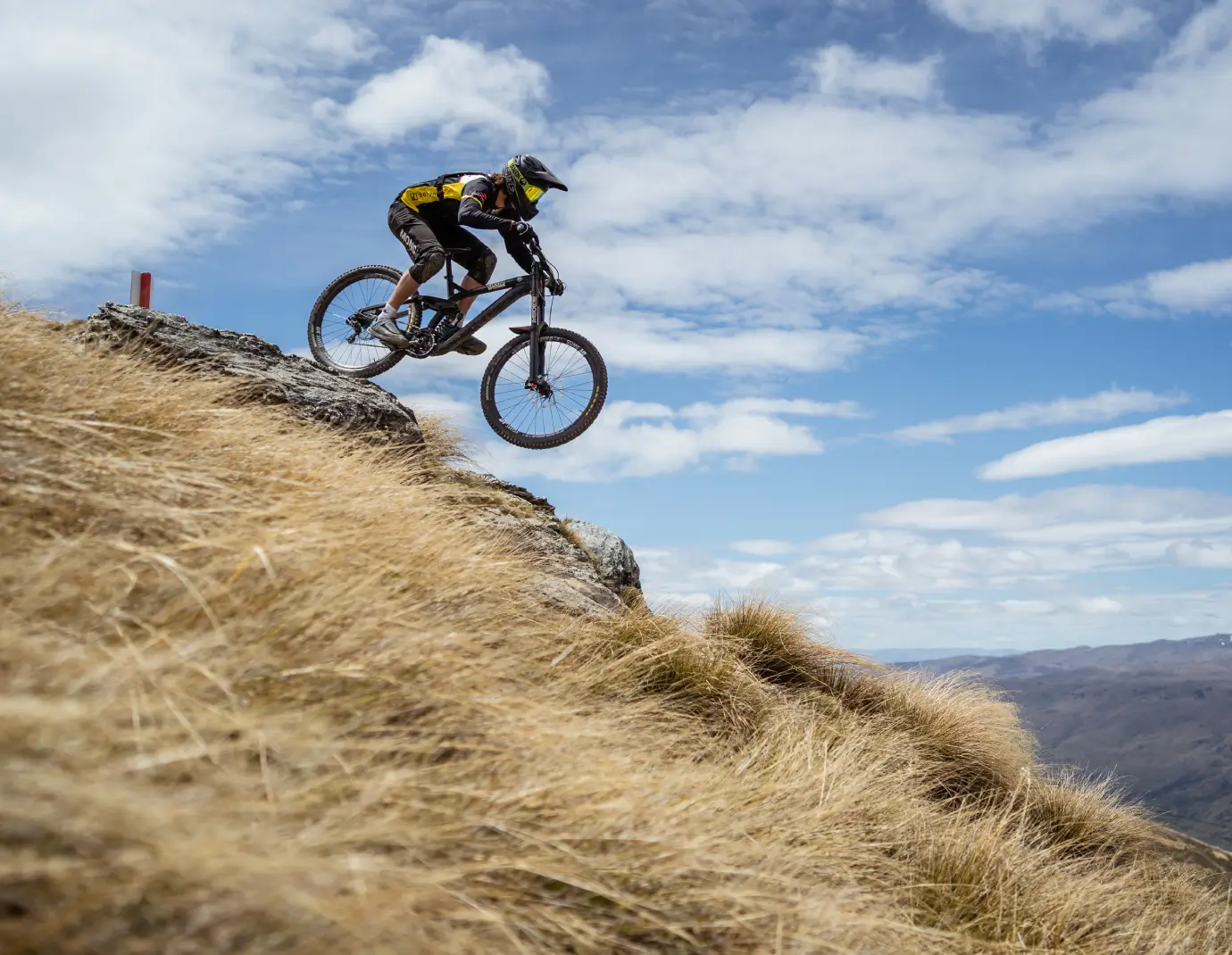 A mountain biker wearing protective gear jumps off a rocky ledge at Cardrona Bike Park, surrounded by golden tussock grass and a scenic alpine landscape.