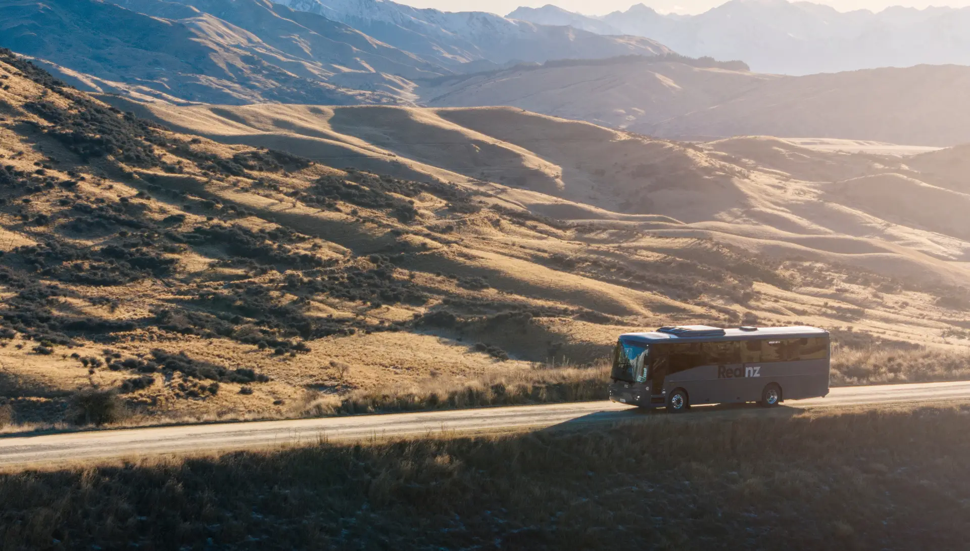 A tour bus travels along a winding road through a golden, mountainous landscape.