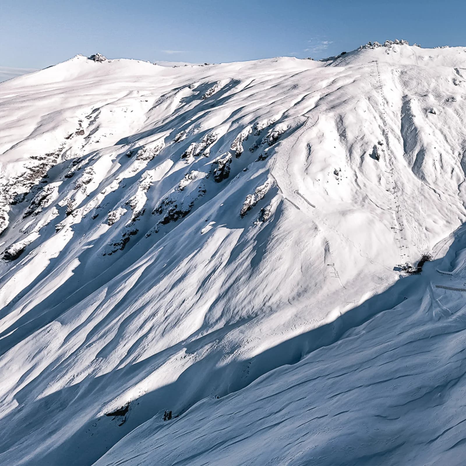 A vast mountain slope blanketed in smooth, untouched snow. The terrain features deep grooves, ridges, and rock outcroppings that create a sense of depth and texture across the mountainside. The high-altitude perspective shows the expansive, open snowfield extending towards the horizon under a clear blue sky. Faint traces of ski tracks and snow fences are visible, indicating human presence while maintaining the natural beauty of the backcountry environment.