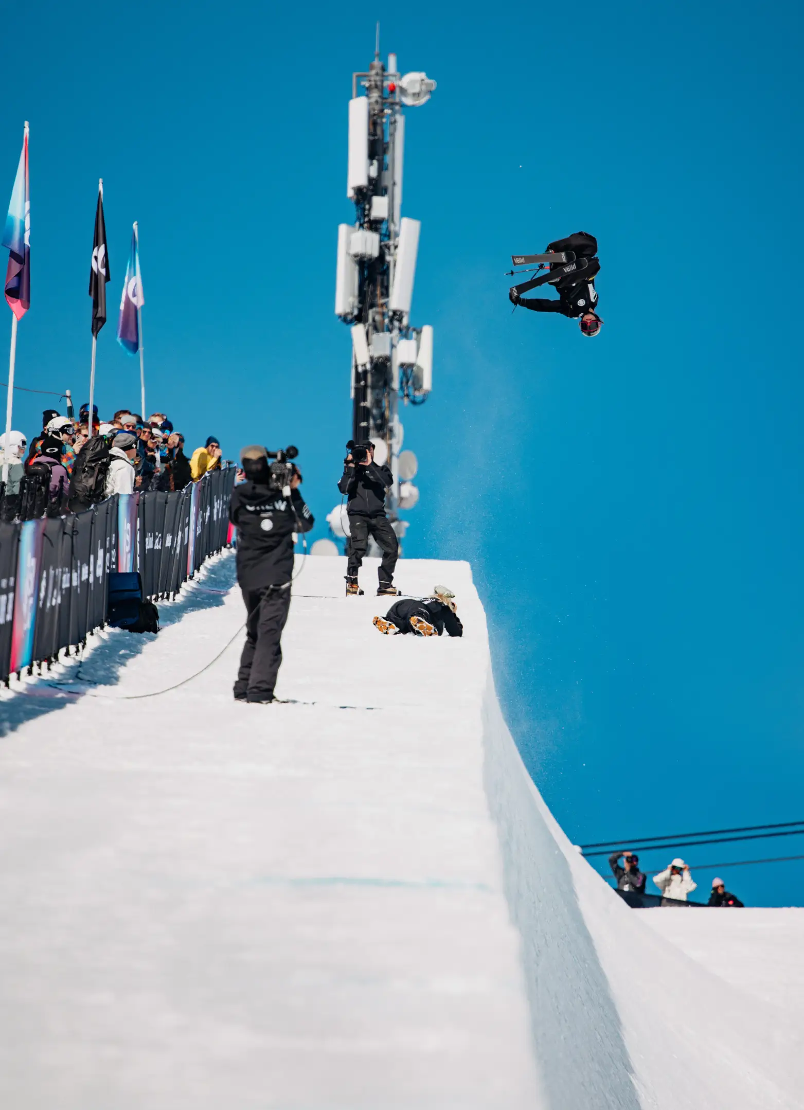 A freestyle skier performs an inverted aerial trick above a snowy competition course as spectators and photographers watch.