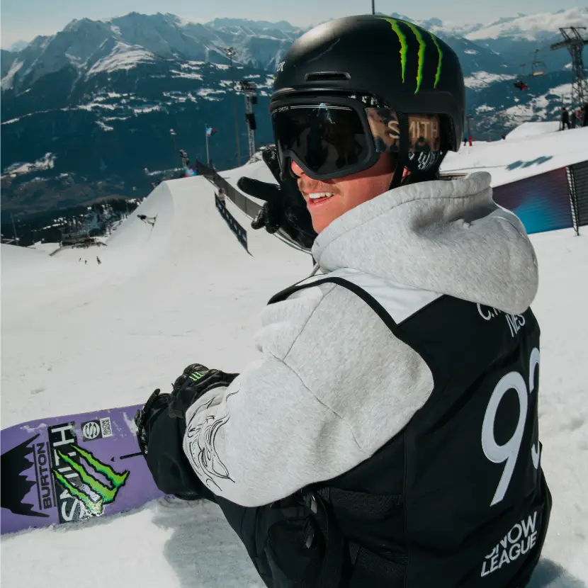 A snowboarder sitting at the top of a slope, wearing a competitor bib.