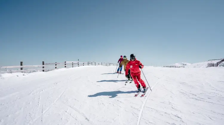 Three skiers on a snow-covered slope, following one another in a line. The skier in the front is dressed in a bright red outfit, while the two behind are in more muted colors. They are all wearing helmets and skiing under a clear blue sky, with a wooden fence lining the left side of the slope.