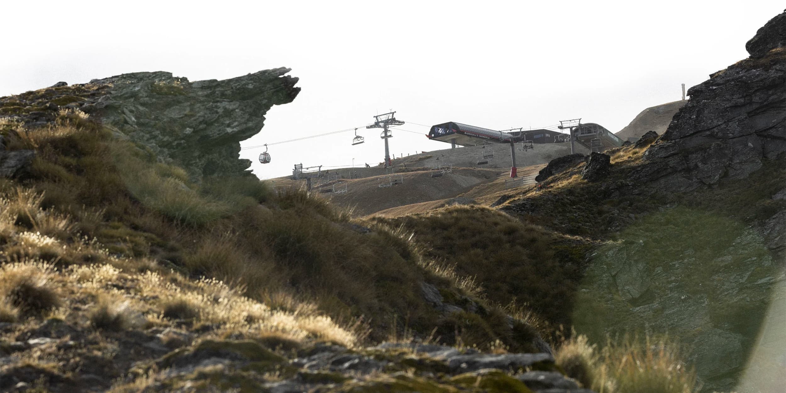 A rugged mountainous landscape with steep rock formations and grassy slopes during summer. A chair lift, with gondolas and chairlifts, is visible in the background.