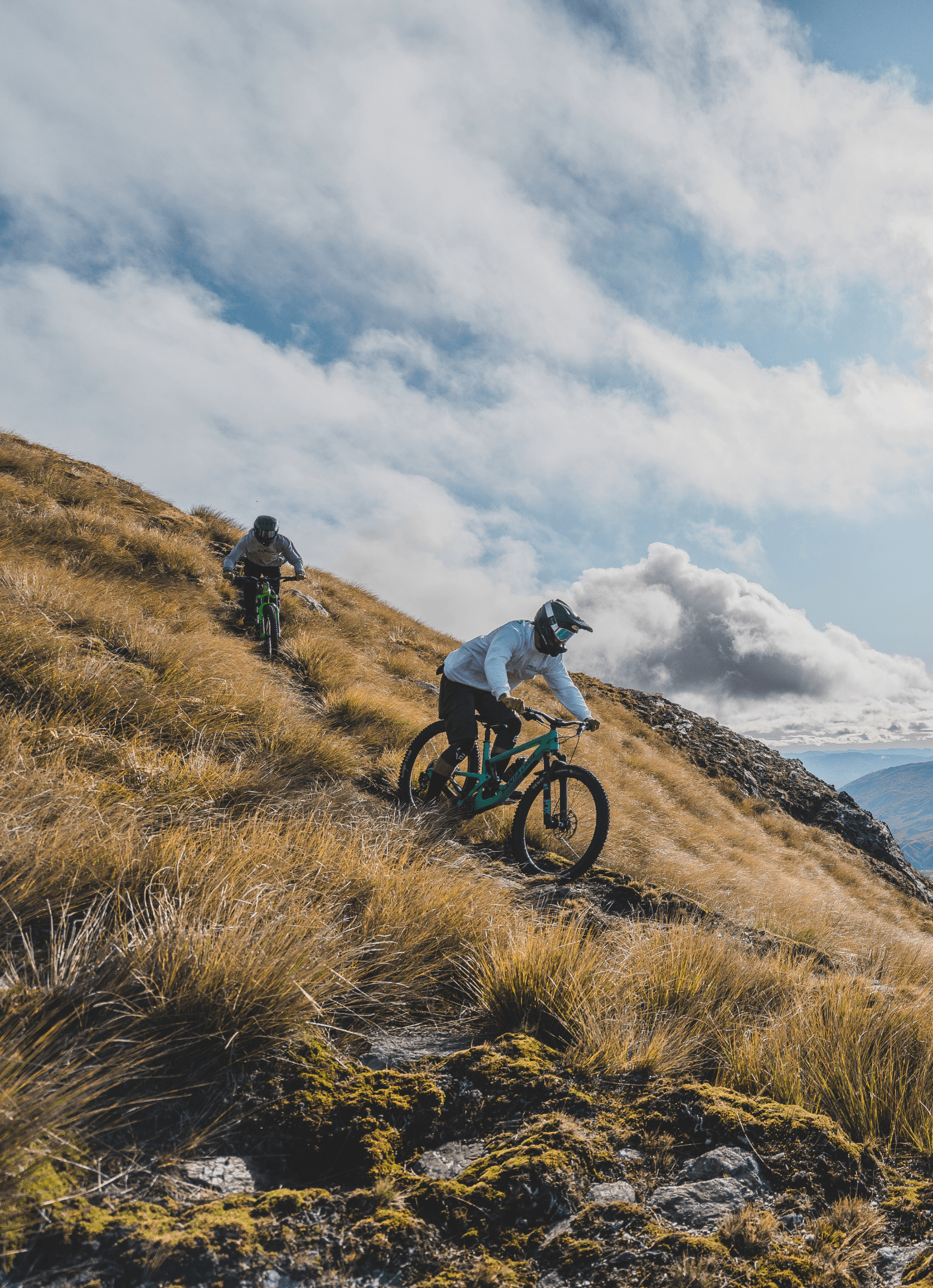 Two mountain bikers descending a grassy hill in full face helmets.