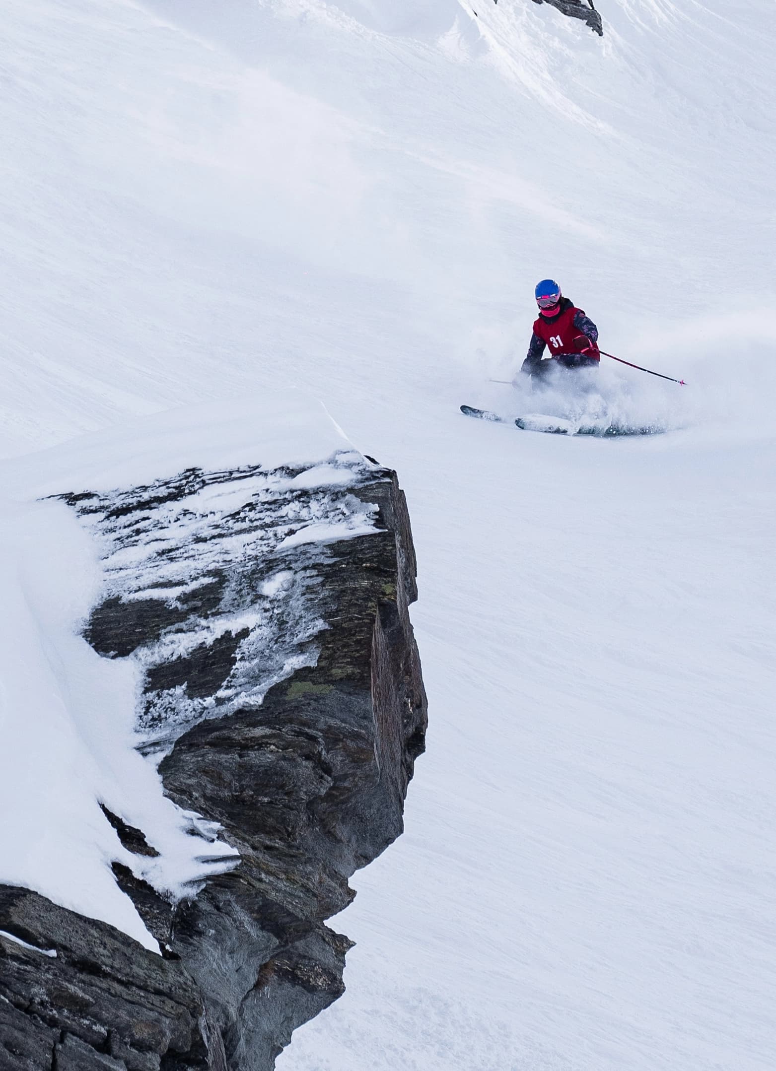 Young skier carving fresh snow near a rock face in a junior freeride competition.