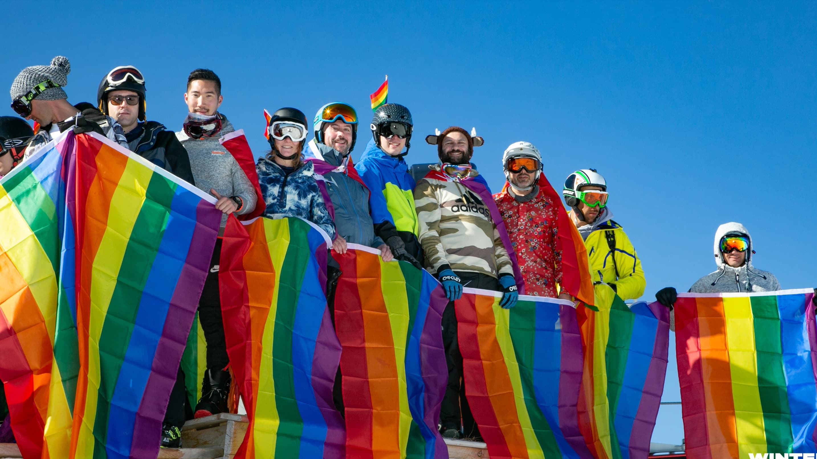 Group of rainbow-flag-waving skiers and snowboarders celebrating Pride on the mountain.