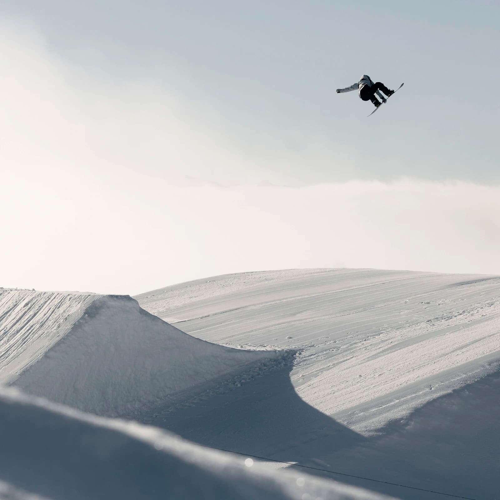 A skier performing an aerial trick above a snowy ramp in a terrain park. The skier is captured in mid-air, soaring against a soft, overcast sky that blends with the expansive, snow-covered landscape below.