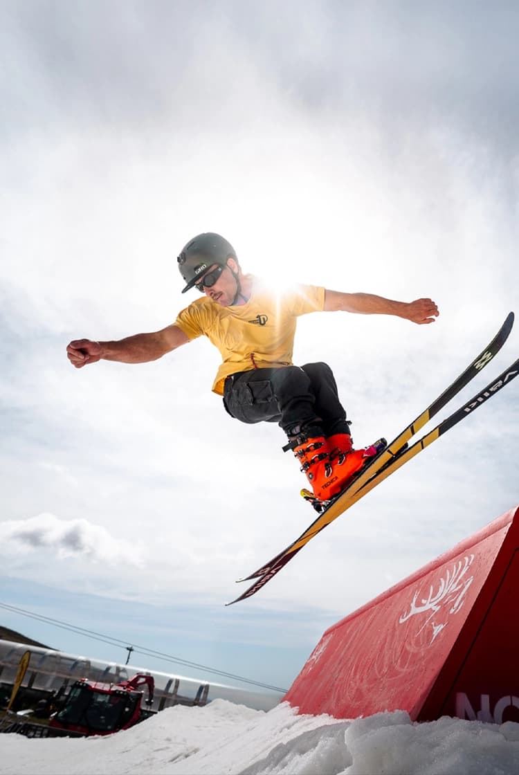 Skier in a helmet and yellow shirt launches into the air above a red terrain-park feature, with bright sun and blue sky behind.