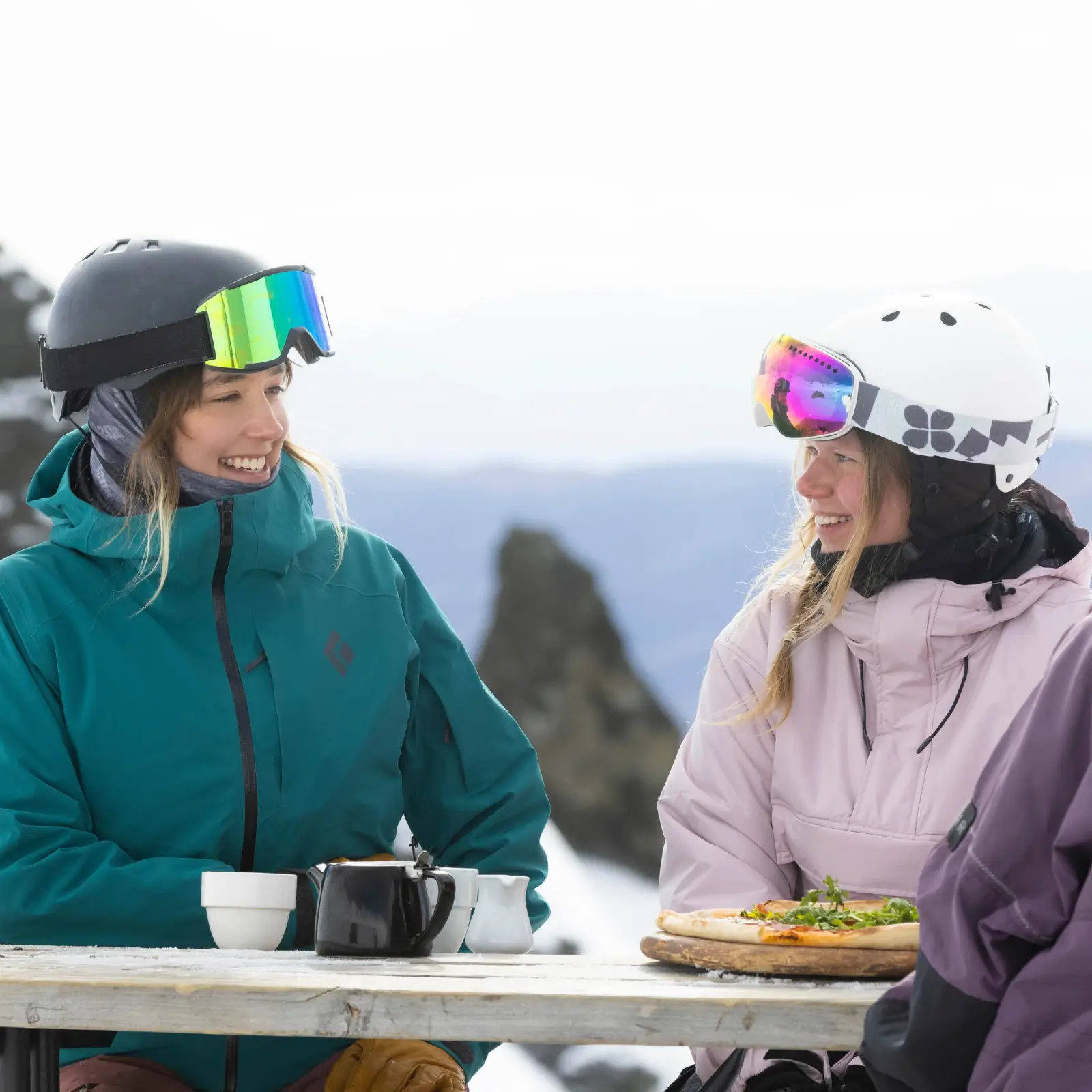 Two skiers in helmets and goggles enjoy hot drinks and pizza at a mountaintop café at Cardrona Alpine Resort, sharing a post-ski moment.