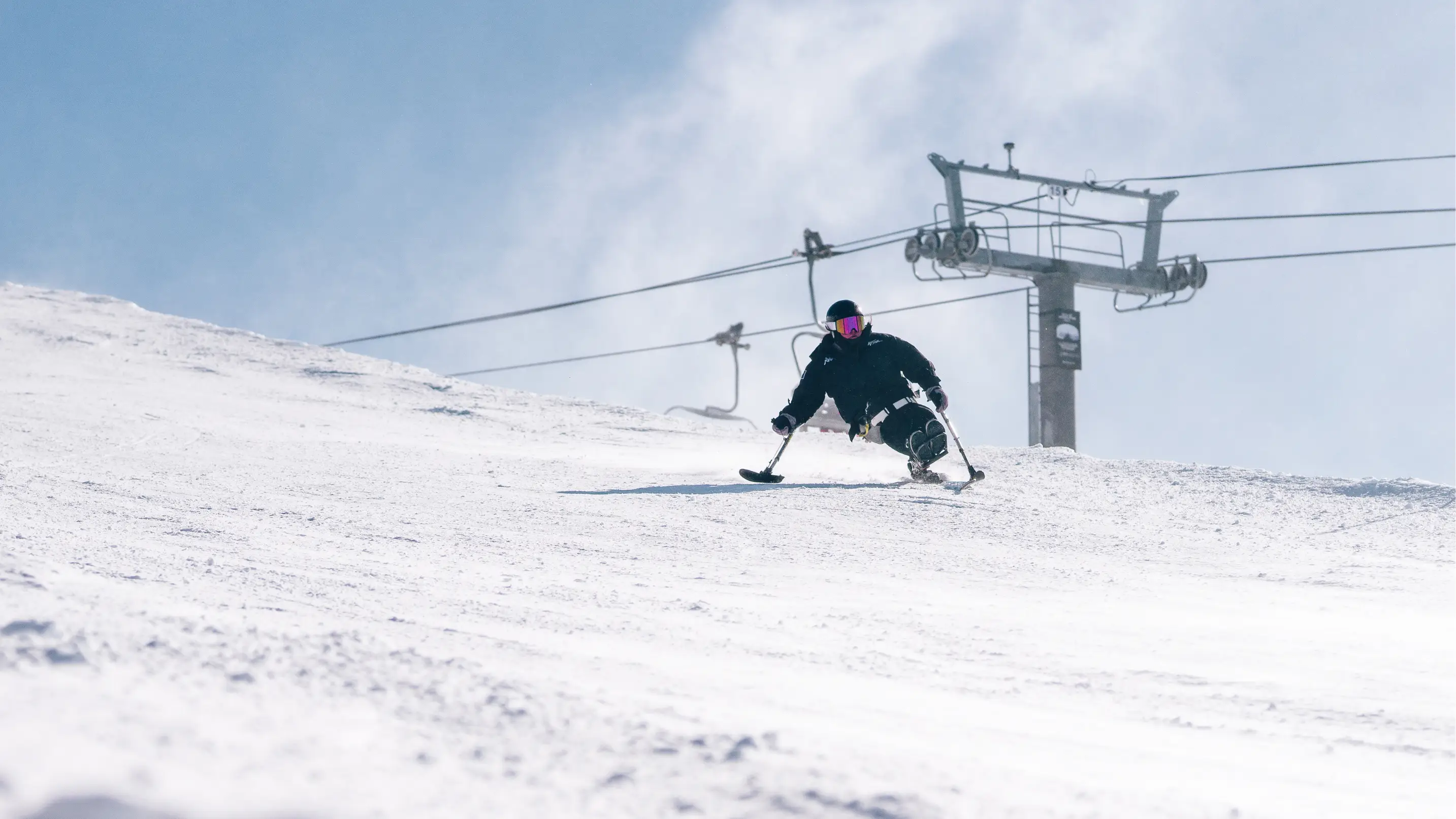 Adaptive sit-skier carving down a snowy slope with a chairlift in the background on a bright winter day.