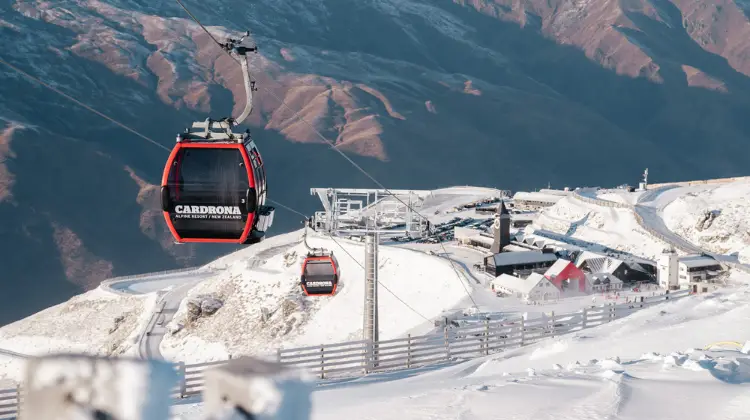 A red Cardrona branded gondola travels up the mountain from the base building on a snowy day.