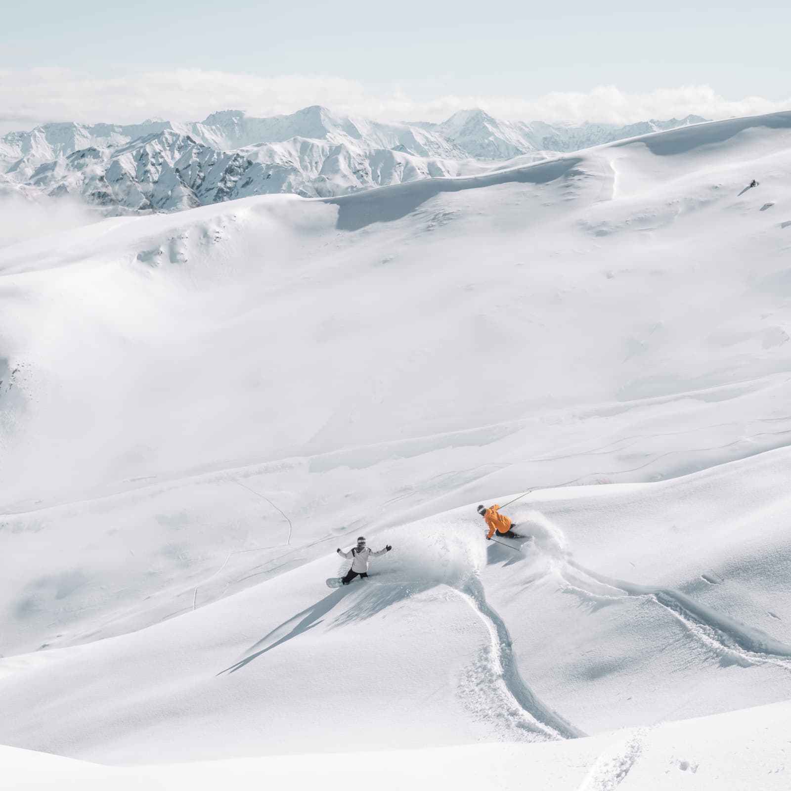 Two skiers carve through fresh powder across a vast snow-covered mountain landscape.