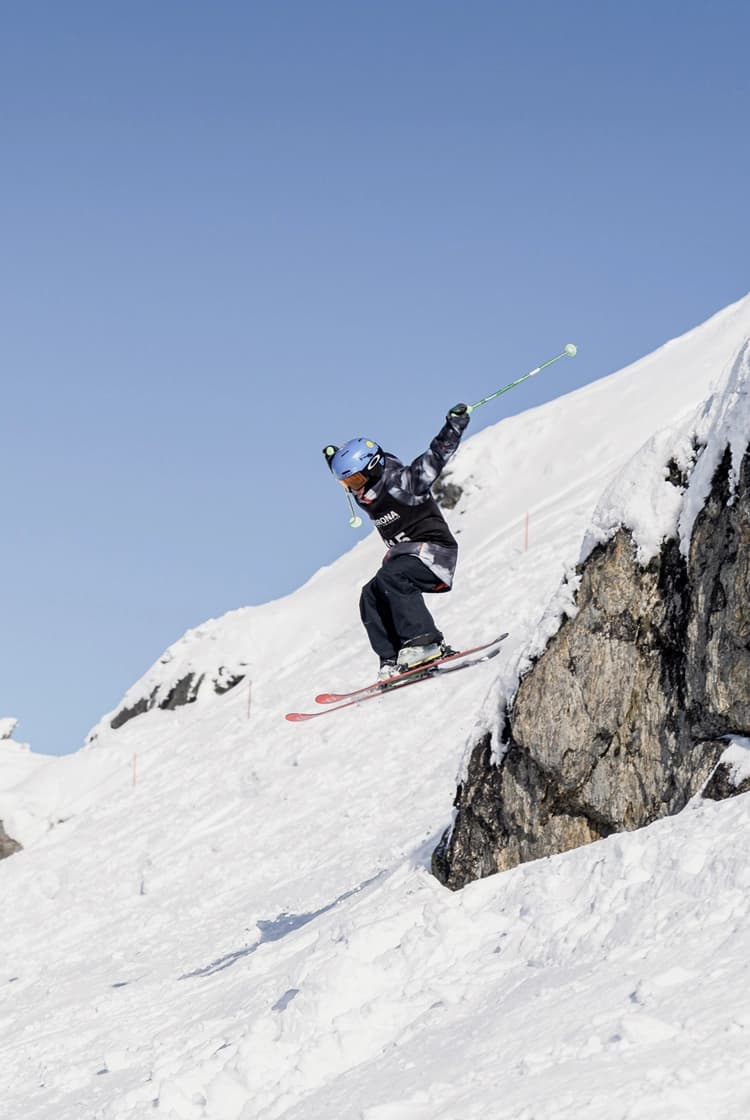 A skier in mid-air launches off a cliff edge against a bright blue sky.