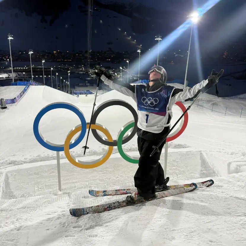 A skier stands in front of the Olympic rings on a snowy slope and celebrated with his arms up in the air.